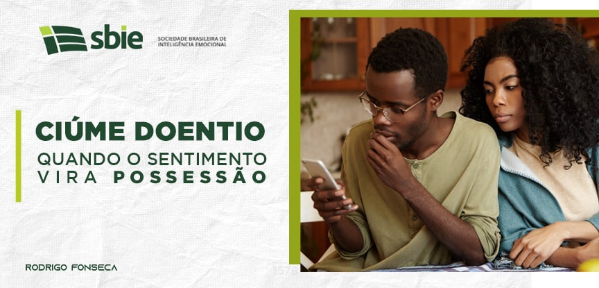 Na imagem, um homem está olhando atentamente um celular em sua mão direita, e atrás dele, está uma mulher com ciúme que, sem que ele perceba, tenta espionar o que ele está fazendo no celular.