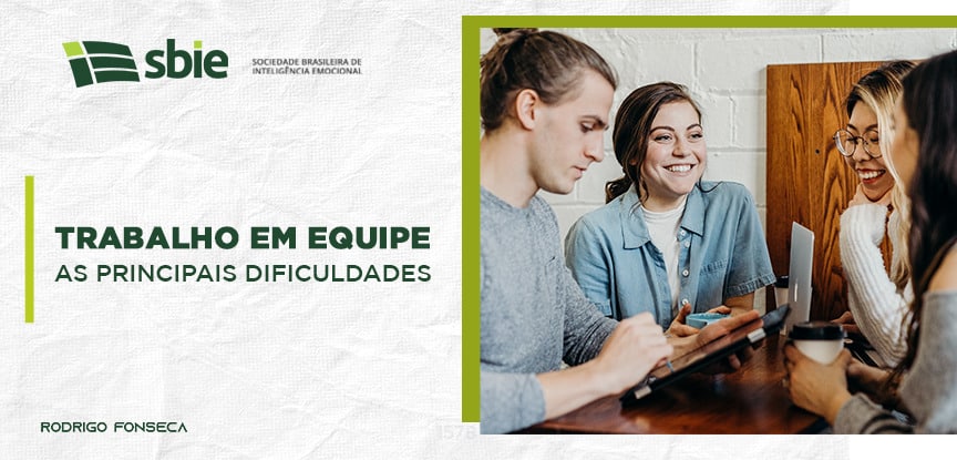 Um grupo de quatro pessoas está reunido e conversando em volta de uma mesa. Eles tem em mãos, um tablet, um copo de café, uma xícara e um notbook. Ao lado está o título: Trabalho em equipe: as principais dificuldades.